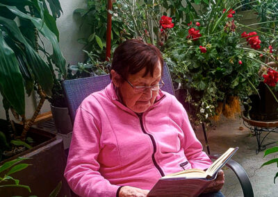An elderly woman is peacefully reading a book in the retirement home greenhouse.